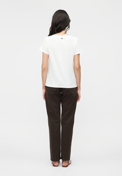 Femme debout de dos, portant un t-shirt blanc uni à manches courtes et un pantalon droit marron foncé avec des sandales.