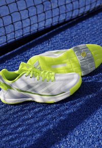 White and neon yellow sports shoes on a blue textured court surface near a black net.