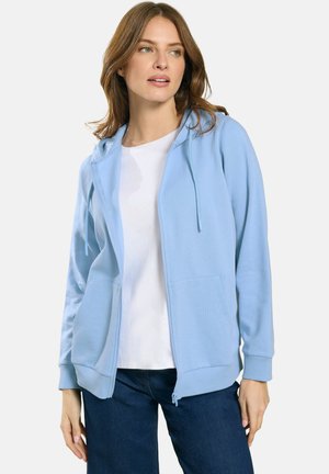 Vrouw draagt een lichtblauwe hoodie met rits over een wit shirt en donkerblauwe spijkerbroek, kijkt iets naar de zijkant tegen een effen achtergrond.