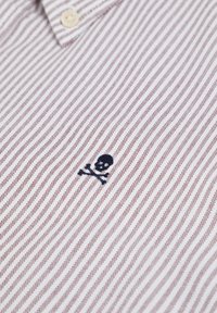 Camisa de algodón con un patrón a rayas rojas y blancas, un cuello con botones y un pequeño bordado de una calavera y huesos cruzados en azul marino.