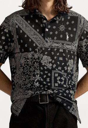 Osoba má na sobě černé polotričko s bílým propracovaným paisley a vzory ve stylu bandany, zastrčené do černých kalhot s černým páskem.