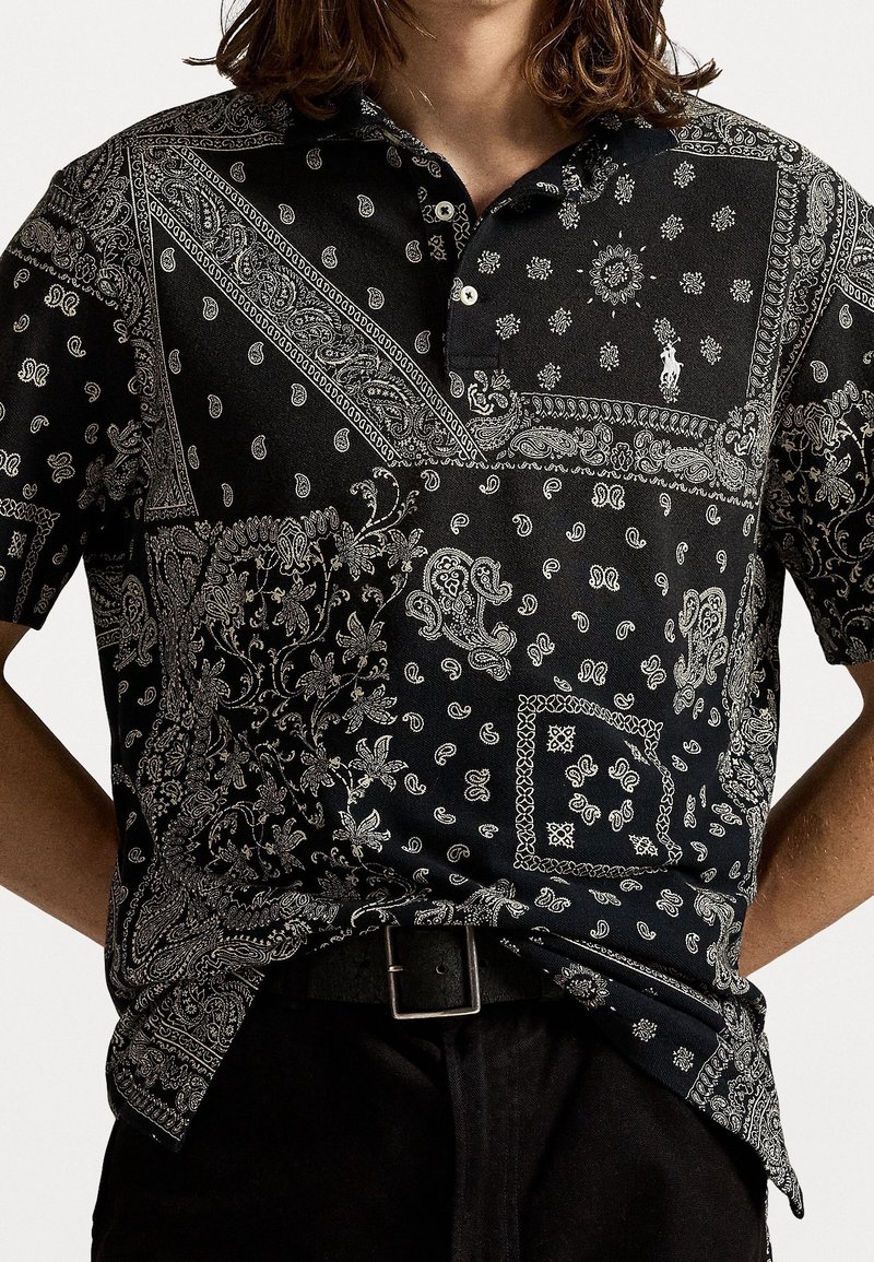 Personne portant un polo noir avec des motifs paisley et style bandana blancs et complexes, rentré dans un pantalon noir avec une ceinture noire.