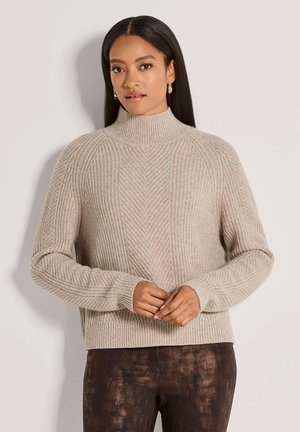 Femme aux longs cheveux foncés portant un pull à col roulé côtelé beige et un pantalon marron foncé texturé, debout contre un mur clair.