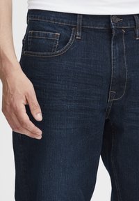 Jeans de mezclilla azul oscuro con corte recto, que presentan un diseño de cinco bolsillos y costuras amarillas en contraste. Textura suave.