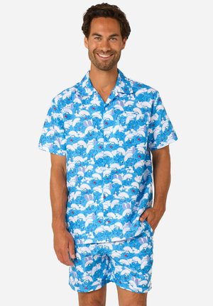 Lachende man die een bijpassend blauw shirt met korte mouwen en korte broek met een cartoon patroon van blauwe Smurfen draagt, staand met één hand in de zak.