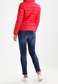 Czerwona kurtka puchowa z wysokim kołnierzem, z pikowaną konstrukcją, w połączeniu z ciemnoniebieskimi rurkami i białymi trampkami.