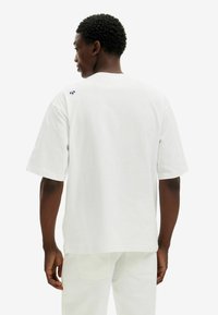 Camiseta blanca de manga corta hecha de algodón; cuenta con un corte holgado y un pequeño detalle de logo en el hombro de la parte posterior. Textura suave.