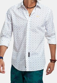 Camisa blanca con lunares azules, mangas largas arremangadas hasta los codos y un pequeño logo en el pecho. La tela parece ligera y con textura.