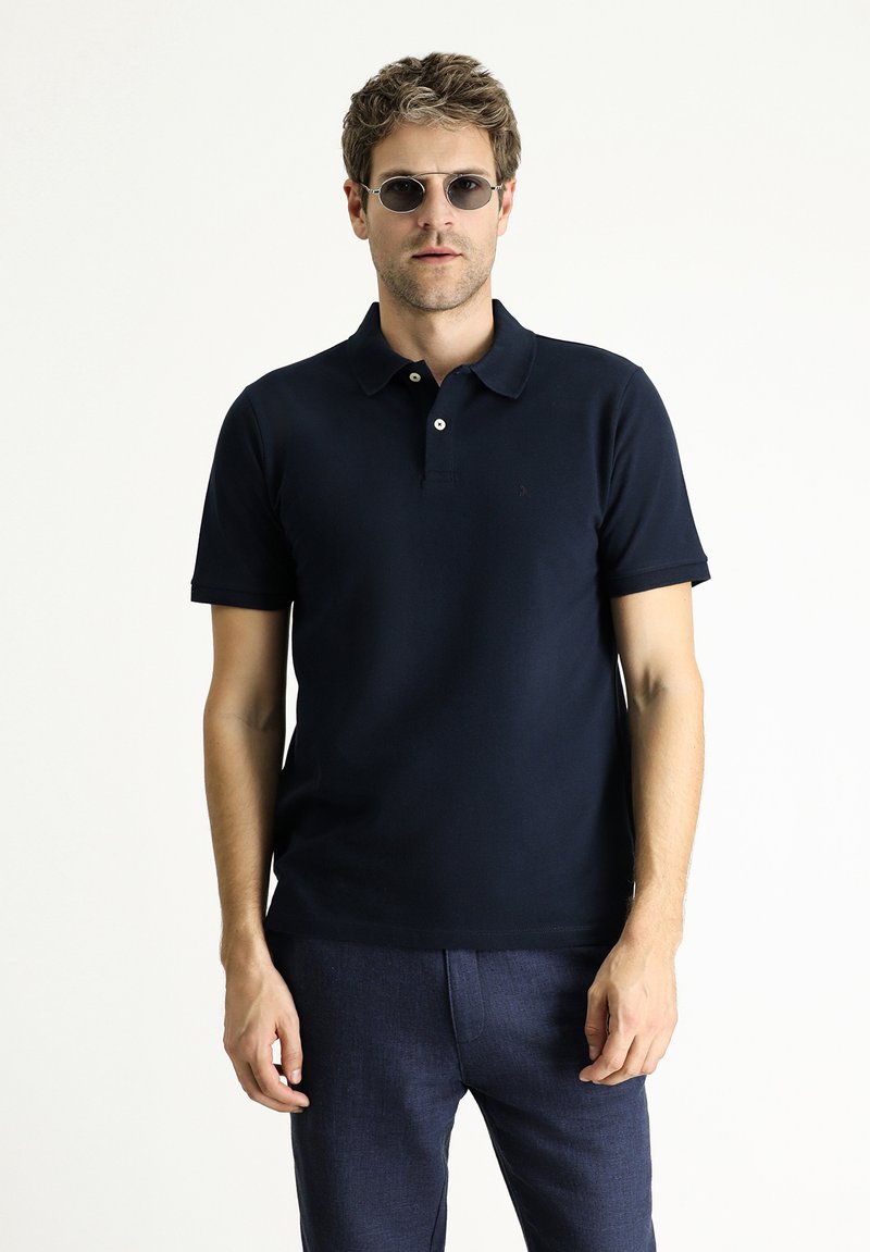 Polo shirt blu navy con maniche corte, patta con due bottoni e tessuto testurizzato. Indossato con pantaloni scuri e occhiali da sole.