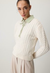 Neulepaita kermansävyssä, jossa on kaapelitekstuuri, ja siinä on vaaleanvihreä kaulus ja nauha. Muotoilu on istuva vyötärö ja ribbaatut hihansuut.