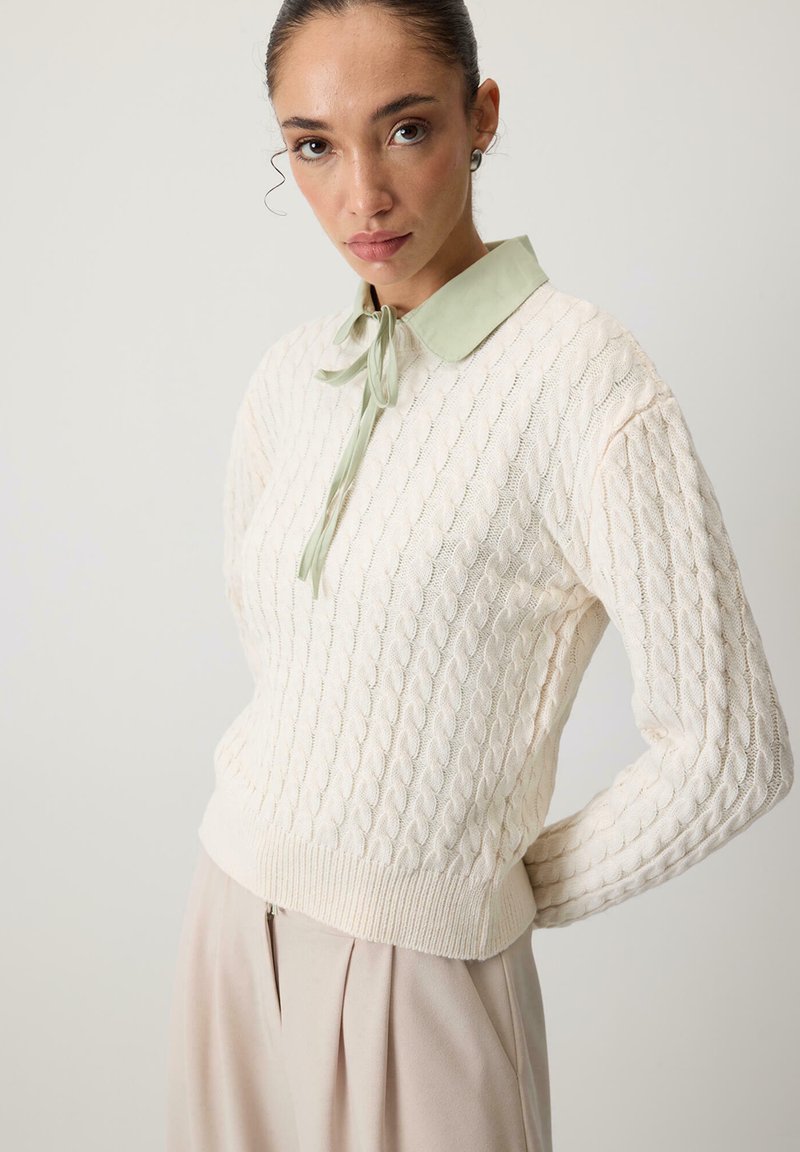 Neulepaita kermansävyssä, jossa on kaapelitekstuuri, ja siinä on vaaleanvihreä kaulus ja nauha. Muotoilu on istuva vyötärö ja ribbaatut hihansuut.