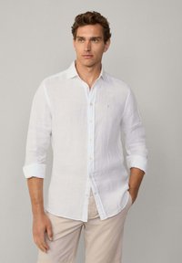 White linen shirt with long sleeves rolled up, featuring a button-down front and a small blue logo on the chest. Smooth texture.