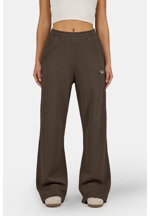 Vrouw met losse bruine geribbelde broek en beige schoenen, staand tegen een effen witte achtergrond.