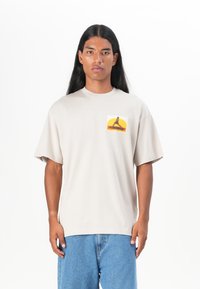 Beige t-skjorte med korte ermer laget av bomull, med en fargerik grafisk trykk på venstre brystlommeområde, kombinert med lyseblå jeans.