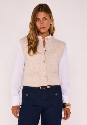Donna che indossa un gilet beige a maglia con bottoni sopra una camicia bianca a maniche lunghe, pantaloni blu scuro, orecchini dorati e diversi braccialetti al polso destro.