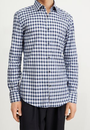 Mężczyzna w koszuli na guziki z długim rękawem w niebiesko-białą kratę, w czarnych spodniach, stojący na tle jednolitego tła.