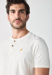 Camiseta blanca de manga corta con cuello henley, dos botones y un pequeño bordado de un águila dorada en el pecho. Textura suave.