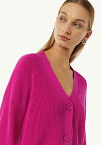 Strickjacke in leuchtendem Pink mit V-Ausschnitt, ausgestattet mit zwei großen runden Knöpfen und geripptem Muster. Weicher Stoff, lockere Passform.