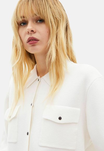 Weißes Hemd mit Knöpfen und Kragen, ausgestattet mit zwei Brusttaschen und schwarzen Knopfakzenten. Der Stoff hat eine strukturierte, gewebte Optik.