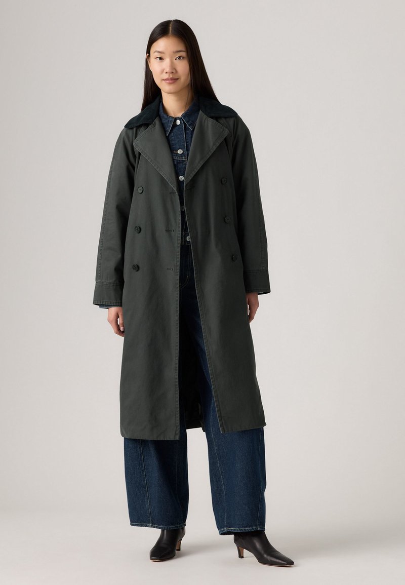 Lang donkergroene trenchcoat met een brede kraag, dubbelgeknoopte ontwerp en subtiele textuur, gecombineerd met high-waisted denim en enkellaarsjes.