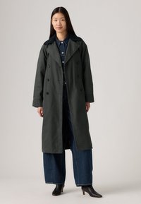 Mørkegrønn trenchcoat med doble knapper, brede slag og lange ermer, kombinert med oversized blå jeans og svarte støvletter med hæl.