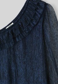 Haut bleu marine avec un col à volants. Fabriqué en un tissu brillant et texturé avec des lignes verticales, il possède des manches transparentes pour une touche supplémentaire.