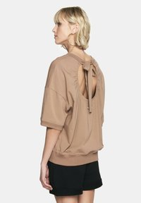 Brązowy oversize'owy sweter z zaokrąglonym wycięciem na plecach, związany kokardką. Krótkie rękawy i miękka tkanina. Noszony z czarnymi szortami.