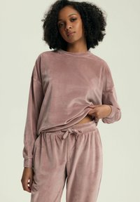 Welurowy zestaw dresowy w miękkim różu, składający się z luźnej długo rękawowej bluzki i spodni z elastycznym pasem oraz sznurkiem ściągającym. Teksturowane wykończenie.
