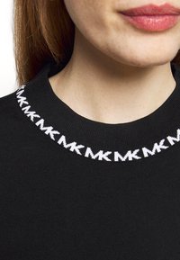 Gros plan sur une personne portant un pull noir avec un motif répété "MK" blanc autour de l'encolure, montrant les lèvres et le cou.