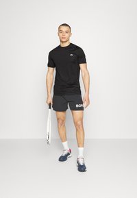 Maglietta sportiva nera a maniche corte con un piccolo logo, abbinata a pantaloni corti grigi scuri con la parola "BORG". Calzini sportivi bianchi e sneakers.