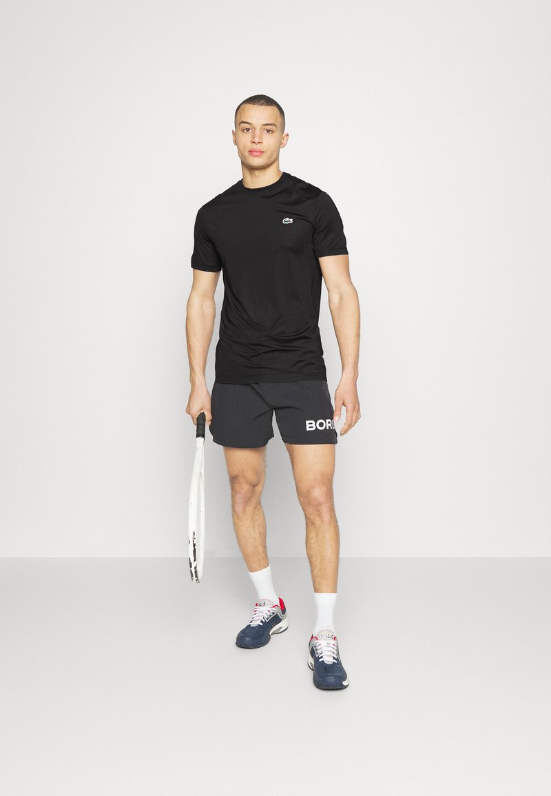 Schwarzes Sport-T-Shirt mit kurzen Ärmeln und kleinem Logo, kombiniert mit dunkelgrauen Shorts, auf denen das Wort "BORG" steht. Weiße Sportsocken und Sneakers.