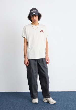 Camiseta blanca de algodón con logo rojo de Nike SB, pantalones cargo grises con bolsillos y una gorra negra con texto blanco, combinados con zapatillas beige.