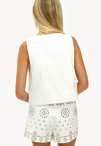 Mujer con cabello largo y rubio, vestida con una blusa sin mangas blanca y pantalones cortos blancos decorados con tachuelas geométricas plateadas, vista desde la parte de atrás.