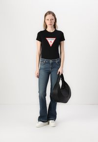 Tricou negru ajustat cu logo triunghiular roșu, purtat cu blugi evazați albaștri. Modelul ține o geantă de umăr din piele neagră și poartă adidași albi.