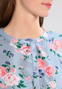 Camicia floreale in tessuto azzurro chiaro con pois bianchi. Presenta fiori rosa e viola, scollatura rotonda e bottoni blu lungo il davanti.