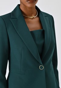 Blazer turchese con rever a punta, dotato di un singolo bottone dorato e tasche laterali discrete. Tessuto liscio con una vestibilità su misura.