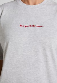 Κοντινό πλάνο ενός ανοιχτογκρι μπλουζακιού με κόκκινο κεντημένο κείμενο που γράφει "σ' αγαπώ μέχρι το φεγγάρι" στο στήθος.