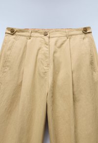 Beige High-Waist-Hose mit seitlichen Verstellern mit Knöpfen, Gürtelschlaufen, vorderen Bügelfalten und seitlichen Taschen vor hellem Hintergrund.