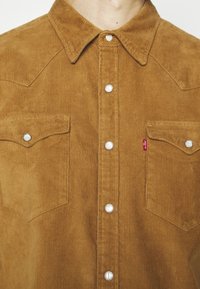 Gros plan sur une chemise en velours côtelé marron avec des boutons-pression blancs et deux poches poitrine, dont l'une comporte une petite étiquette rouge Levi's.