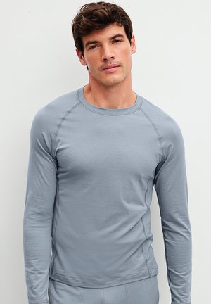 Man met een lichtblauw overhemd met lange mouwen en bijpassende broek, staand tegen een effen witte achtergrond, kijkend naar de camera met een neutrale uitdrukking.