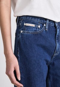 Tamsiai mėlynos džinsinės kelnės su aukštu liemeniu, įskaitant raudonai baltą Calvin Klein logotipo etiketę ir sidabrinius priedus.