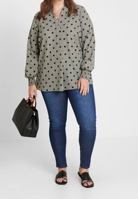 Blouse grise à pois avec des manches bouffantes, dotée d'un col en V, associée à un jean skinny bleu foncé et des sandales noires. Comprend un sac à main noir.