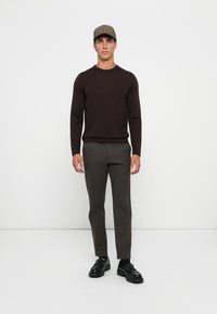 Dark brown long-sleeve sweater paired with tailored gray pants and black shoes. Model wearing a green checkered cap. Solid colors, fitted design.