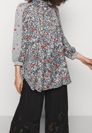 Femme portant un chemisier à col montant avec des fleurs multicolores et des manches bouffantes, associé à un pantalon noir à jambes larges avec des découpes.