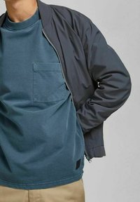 Grå bomberjacka med ribbade manschetter över en teal crewneck t-shirt med bröstficka. Avslappnad passform med en mjuk textur.