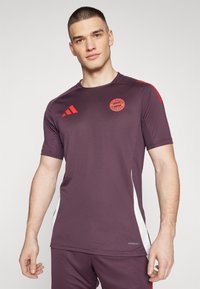 Mörkröd atletisk tröja med röda accenter och vita sidopaneler, med FC Bayern Münchens logotyp och lätt, ventilerande tyg.