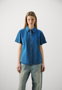 Kortermet denimskjorte i mellomblå farge, med knappelukking foran, spiss krage, og avslappet passform med litt avrundede kanter.