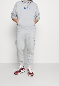 Světle šedá mikina s modrým logem Nike, ladící šedé tepláky s cargo kapsami a červené a bílé tenisky Nike s texturovanou podrážkou.