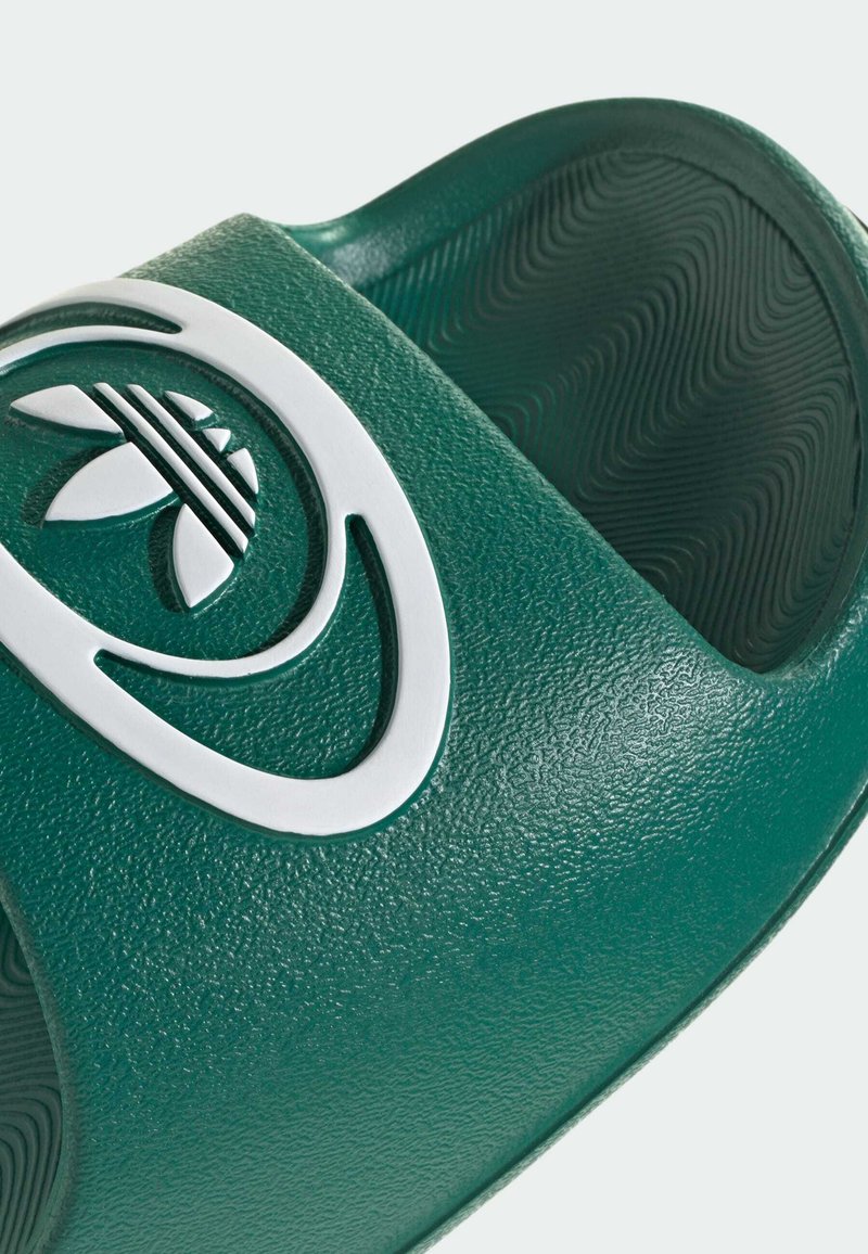 Grøn slip på sandal med tekstureret materiale, bred rem og fremtrædende hvid logo-detalje. Sålen har et bølgemønster for bedre greb.