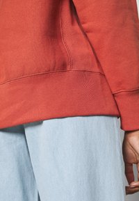 Czerwony sweter w odcieniu rdzy z teksturowanej tkaniny, z prążkowanymi mankietami i dołem, zestawiony z jasnoniebieskimi spodniami dżinsowymi. Dodatki to widoczne szwy.